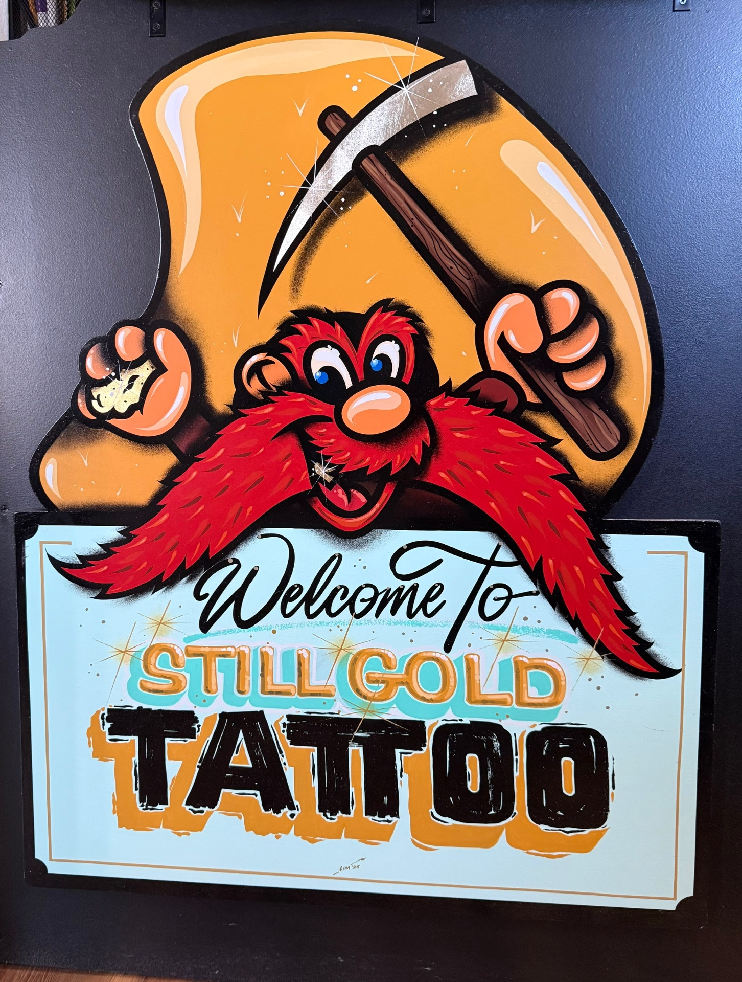 Hand-painted Welcome to Still Gold Tattoo sign with gold miner mascot